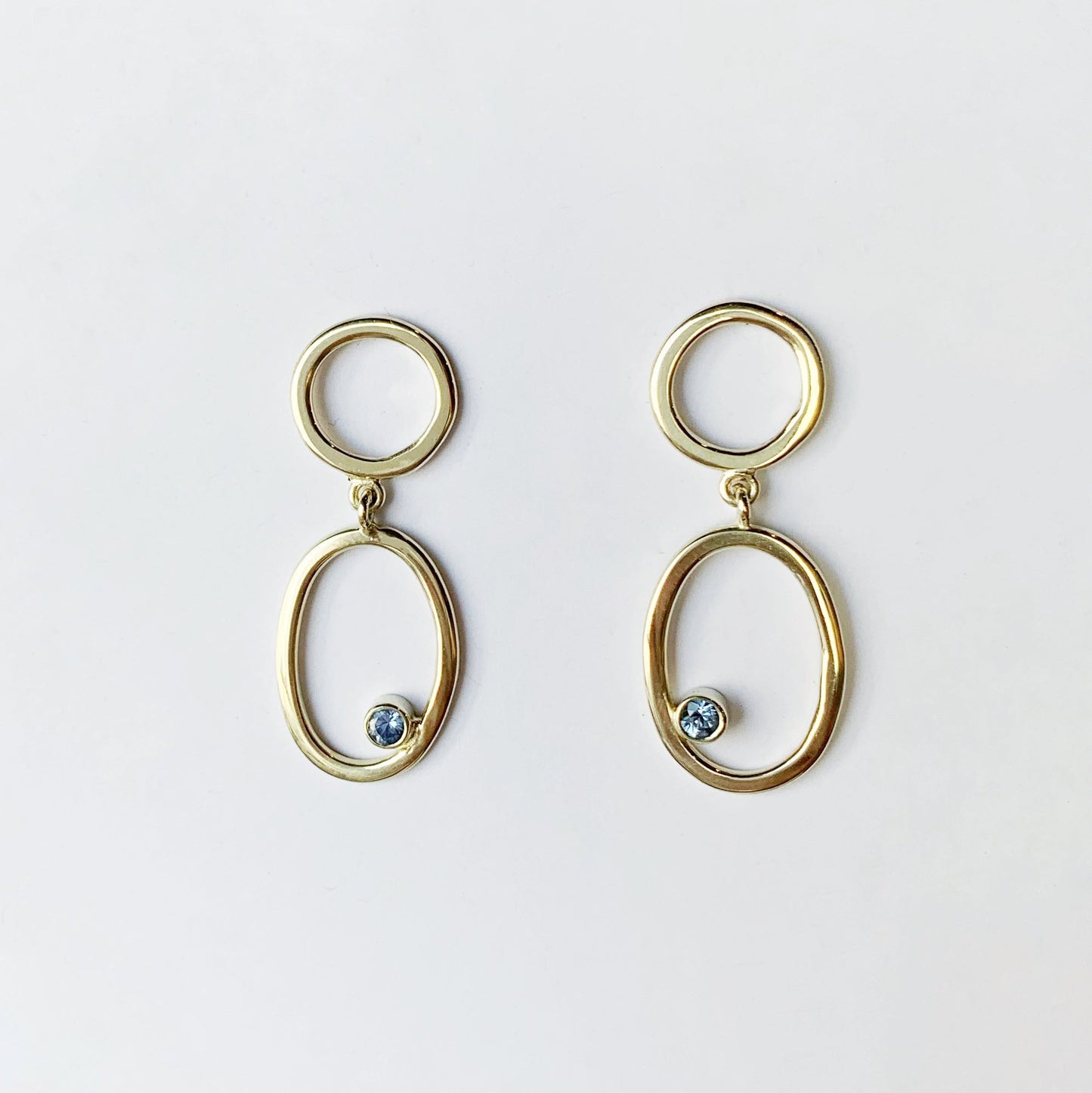 Productfoto op witte achtergrond van geel gouden oorbellen. Plat op het oor een cirkelsteker met daaronder een ovaal waarin een facet geslepen saffier is gezet