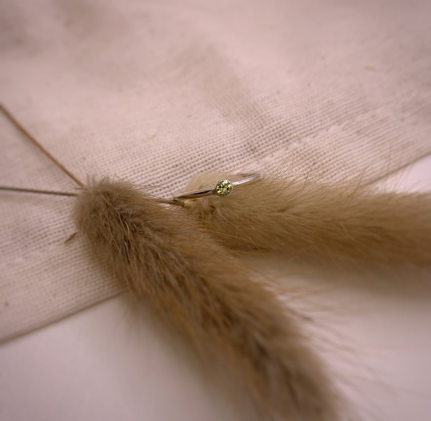 Productfoto op stoffen achtergrond. Zilveren ring met facet geslepen groene peridot in gladomzetting. Zetting is tussen de ring geplaatst.