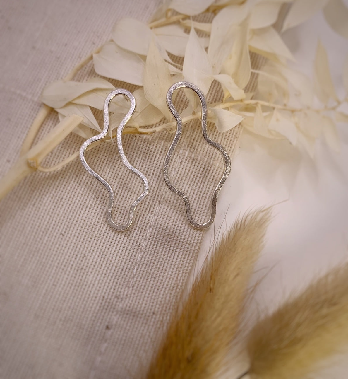 Productfoto op stoffen achtergrond. Asymmetrische oorbellen met vrije vorm. Voor- en achterzijde gematteerd, binnen en buitenkant zijn hoogglans