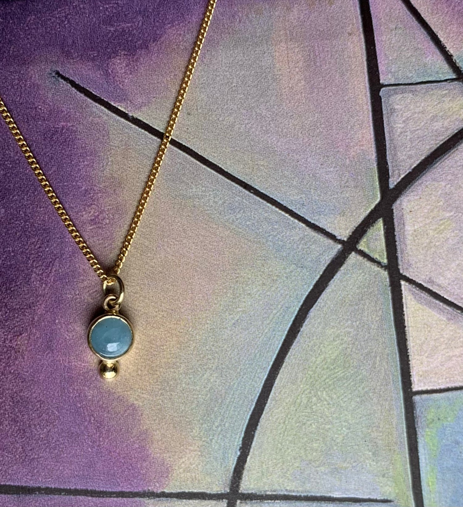 Productfoto op gekleurde achtergrond, close-up van aquamarijn hanger met gouden balletje. Hangt aan een collier met gourmet schakel