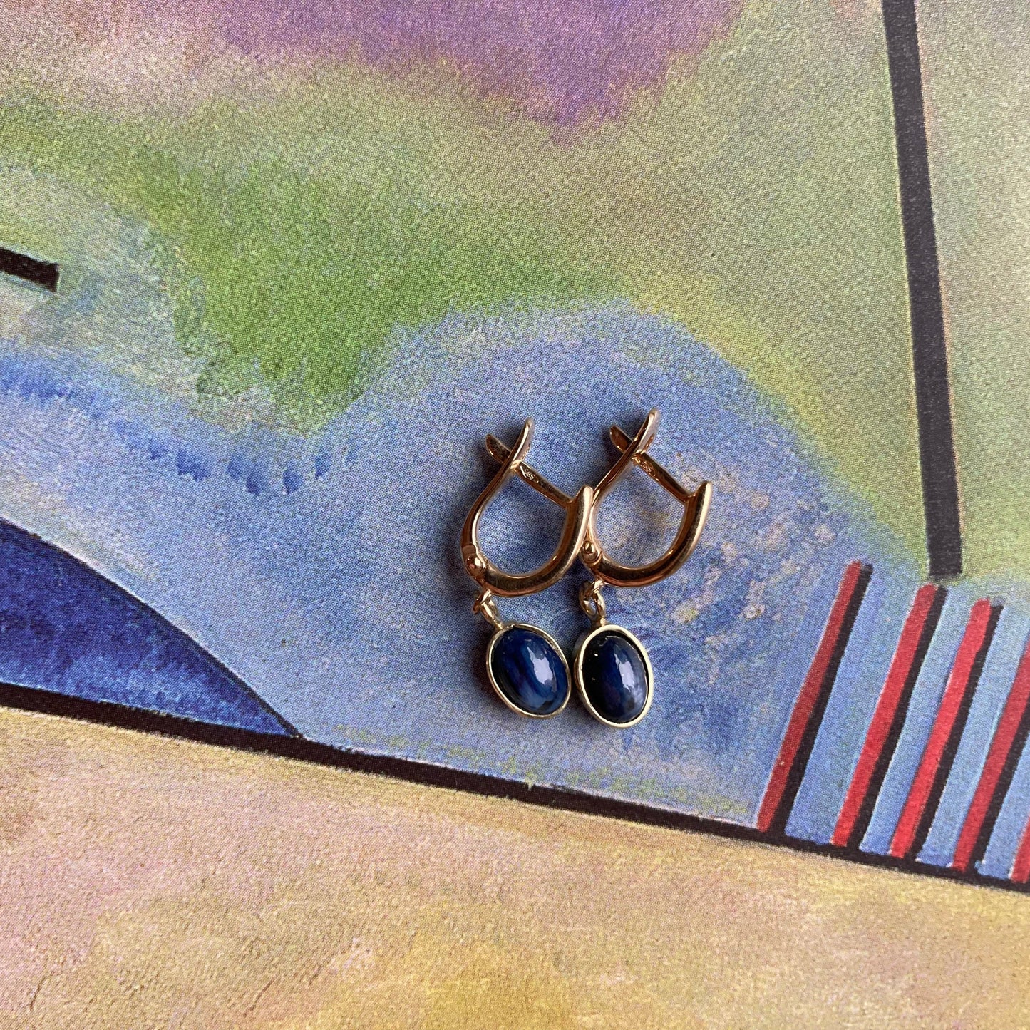 Productfoto op gekleurde achtergrond, Geelgouden "huggie" brisuren met ovaal, cabochon geslepen, donkerblauwe saffieren als bedels