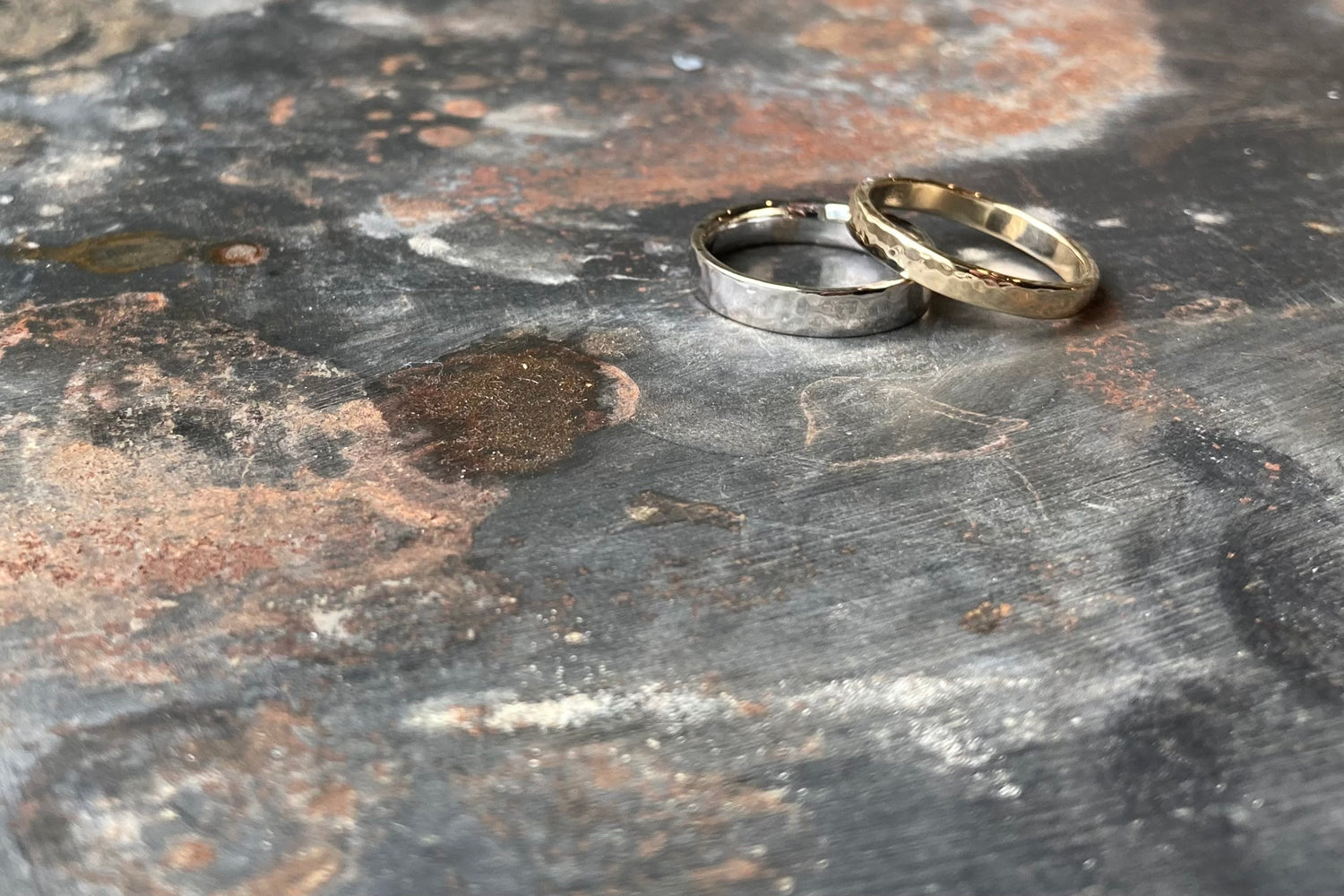 Set trouwringen op grove ondergrond. Een geelgouden ring ligt schuin bovenop een witgouden ring. Beide ringen met hamerslag bewerkt