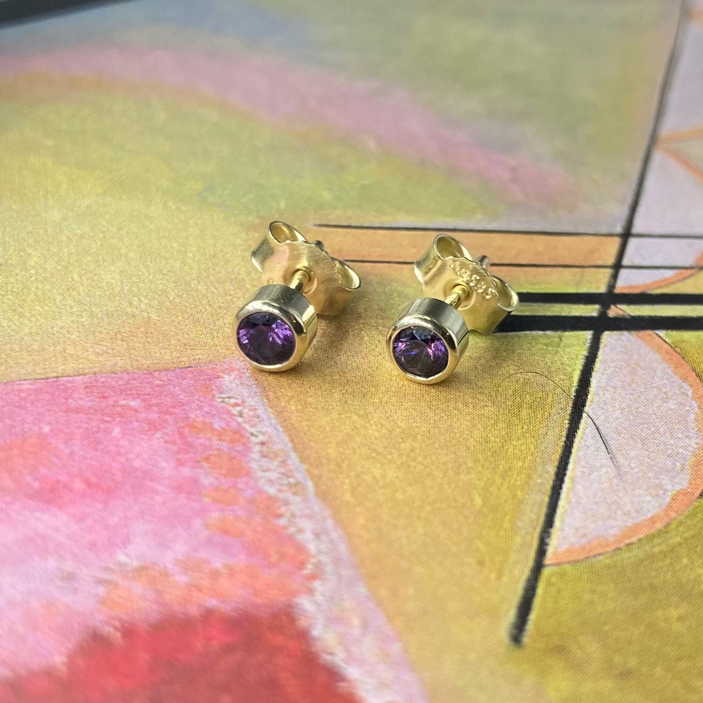 Productfoto op gekleurde ondergrond, geelgouden oorknopjes met facet geslepen paarse granaat